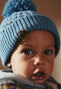 Bonnet en tricot bleu avec un pompon, présentant une texture côtelée et des motifs en losange, porté par un enfant aux cheveux bouclés et aux grands yeux foncés.