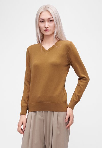 Jeune femme aux longs cheveux blonds portant un pull marron à col en V et un pantalon beige à jambes larges sur un fond clair uni.