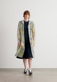 Long hooded coat in a marbled pastel pattern, with elastic drawstring at the waist and lightweight material, over a navy dress and black sneakers.