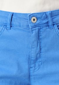 Siniset farkut, joissa on nappikiinnitys, erottuvat sileästä tekstuuristaan ja jämäkästä muotoilustaan. Näkyvät saumat vyötäröllä ja taskuissa.