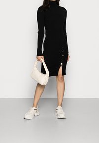 Černé žebrované pletené šaty s dlouhými rukávy, vysokým límcem, zapínání na knoflíky na levé straně, v kombinaci s pletenou bílou kabelkou a teniskami.