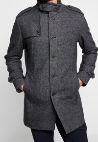 Grå ullkappa med mönstrad textur, knäppning med knappar och design med hög krage. Två framfickor; figursydd silhuett.