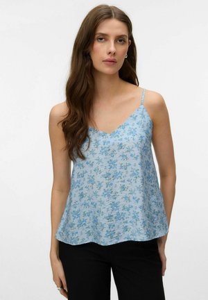 Kvinde med langt brunt hår iført en lyseblå, blomsterprintet ærmeløs top og sorte bukser, stående foran en ensfarvet hvid baggrund.