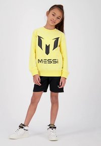 Žlutá mikina s černým grafickým logem "MESSi" na přední straně, kombinovaná s černými kraťasy. Outfit doplňují bílé vysoké tenisky.