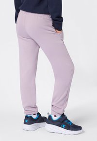 Person i ljuslila sweatpants och marinblå sneakers med vita sulor och blå detaljer, stående med händerna i fickorna.