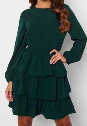 Femme portant une robe verte foncée à manches longues avec un col rond et une jupe à plusieurs couches.