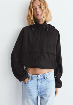 Mladá osoba v čiernej krátkej bunde na zips s kapucňou a svetlomodrých džínsach s vysokým pásom, stojaca s jednou rukou v vrecku.