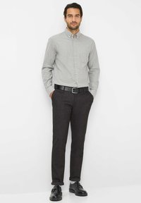 Lichtgrijze knoop shirt met een zak, gecombineerd met donkergrijze gestructureerde broek en een zwarte riem. Het model staat met de handen in de zakken.