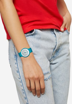 Et turkis silikoneur med en rund hvid urskive og blå markeringer, båret på et håndled, kombineret med en rød skjorte og slidte lyse jeans.