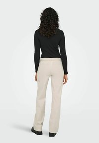 Femme aux cheveux bouclés portant un haut à manches longues noir, un pantalon beige et des chaussures noires, debout de dos sur un fond uni.