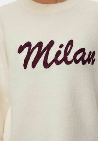 Kremasti pleteni pulover s riječju "Milano" izvezanom preko prsa u tamnoburgundnoj kurzivnoj pisanji.