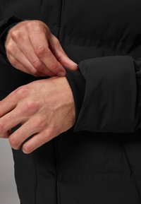 Chaqueta acolchada negra con puños acanalados. Manos ajustando el puño, mostrando las costuras detalladas y la textura acolchada.