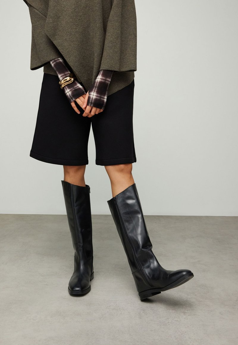 Personne portant des bottes en cuir noires montantes jusqu'aux genoux, un short noir longueur genoux, un haut vert olive et une chemise à carreaux à manches longues avec un bracelet en or.
