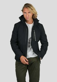 Veste noire avec un col haut, fermeture éclair à l'avant et poches latérales ; affiche le logo "REDSKINS" sur la fermeture éclair ; matériau extérieur lisse.