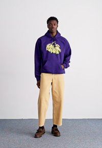 Paarse hoodie met gele grafische ontwerpen, gecombineerd met beige broeken en bruine schoenen. Model staat tegen een effen achtergrond.