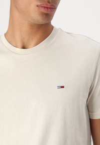 Tricou din bumbac bej cu guler rotund, având un mic logo brodat în nuanțe de bleumarin, roșu și alb pe partea stângă a pieptului. Textură moale.
