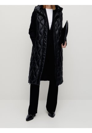 Person iført lang sort quiltet vest over sort outfit, holder en sort clutch, står mod en ensfarvet lys baggrund.