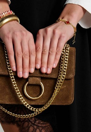 Bruine suède handtas met gouden kettingband, voorzien van een cirkelvormige metalen ringdetail. Handen met een gouden armband en verschillende gestructureerde koordarmbanden rusten op de tas.