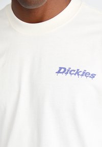 Bjela pamučna majica s logotipom u ljubičastom otjecanju koji glasi "Dickies" na lijevoj strani prsnog koša. Mekana tekstura s klasičnim okruglim izrezom.