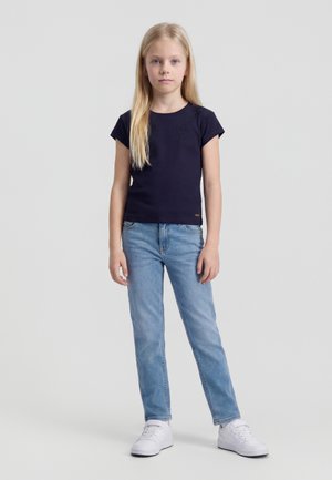 Młoda dziewczyna z długimi blond włosami stoi ubrana w granatową bluzkę z krótkim rękawem, jasnoniebieskie jeansy i białe trampki na jednolitym tle.