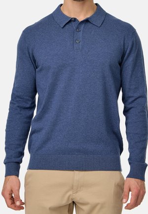 Homme portant un polo en maille bleu à manches longues avec trois boutons, associé à un pantalon beige, devant un fond clair uni.