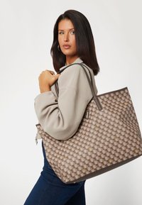 Grand sac fourre-tout avec un motif à damier marron et beige, texture lisse et longues bandoulières. Des accents dorés subtils viennent embellir le design.