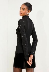 Czarny prążkowany sweter z długimi rękawami, zdobiony perłowymi dodatkami w kolorze srebra. Modelka ma na sobie czarną aksamitną spódnicę, podkreślającą dopasowaną sylwetkę.