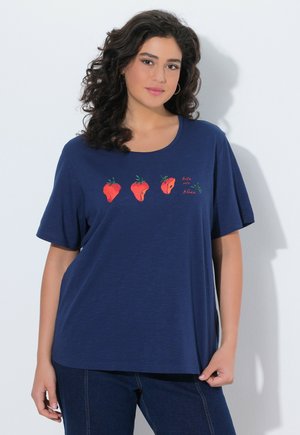 Kvinde iført en mørkeblå t-shirt med tre jordbær og teksten "bite into bliss" trykt i rødt på brystet, kombineret med mørke jeans.