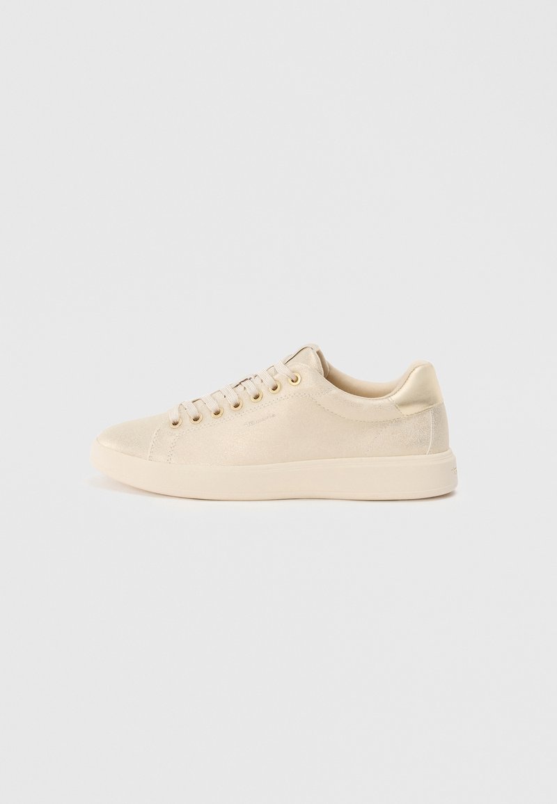 Lave topp beige sneakers med snøring, gulløyer, glanset hælpatch og en glatt gummisåle på en hvit bakgrunn.