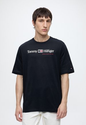 Ung mand med kort mørkt hår iført en sort Tommy Hilfiger t-shirt med hvid tekst og hvide bukser mod en ensfarvet lys baggrund.