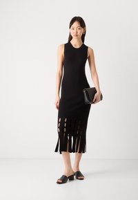 Melnā adīta kleita bez piedurknēm, kurai ir fringu un izgriezumu detaļas apakšmalā. Apvienota ar melniem sandalēm ar slīdēšanu un sajūga somu.