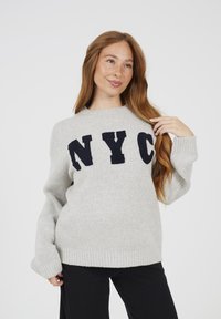 Kobieta z długimi czerwonymi włosami, ubrana w jasnoszary sweter z literami "NYC" i czarne spodnie, pozująca na jednolitym tle.