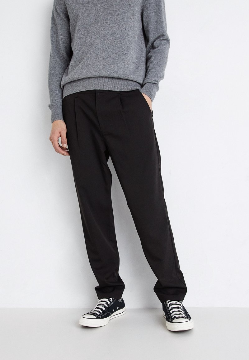 Osoba stoi w czarnych, casualowych spodniach, szarym swetrze i czarno-białych sneakersach, z ręką w kieszeni na białym tle.