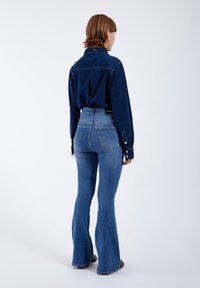 Camisa de mezclilla en azul oscuro con puños abotonados, combinada con jeans de campana claros de cintura alta. Textura suave y diseño ajustado.