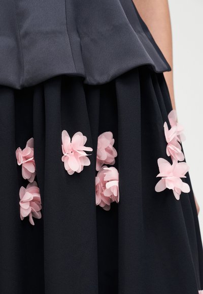 Μαύρη φούστα με πιέτες που διαθέτει στρωματοποιημένα ροζ ανθικά διακοσμητικά στο κάτω μέρος, κατασκευασμένη από ελαφρύ ύφασμα, προσθέτοντας υφή και λεπτομέρεια.