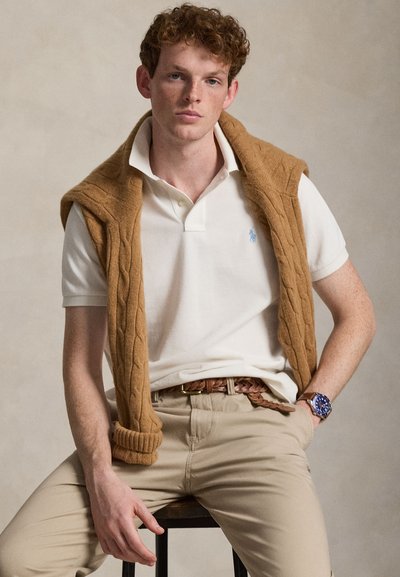 Νεαρός άνδρας με σγουρά μαλλιά που κάθεται σε σκαμπό, φοράει λευκό πόλο, μπεζ πουλόβερ πλεκτό με σχέδιο cable draped στους ώμους, μπεζ παντελόνι και μπλε ρολόι χειρός.