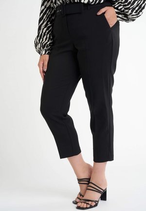 Kobieta ubrana w czarne, skrócone, dopasowane spodnie z paskiem, czarne sandały na obcasie z paskami oraz bluzkę w zebrowy wzór z bufkami na rękawach.