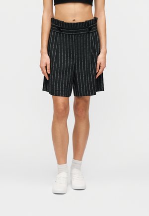 Pantalones cortos a rayas negras con cintura alta, dos botones frontales y corte holgado. Combinados con zapatillas blancas y calcetines acanalados, sobre un fondo liso.