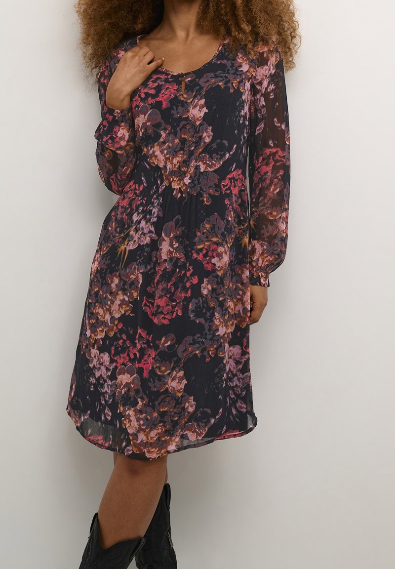 Femme portant une robe fleurie foncée mi-longue avec des manches longues en voile transparent et des bottes de cowboy noires, debout devant un fond clair uni.