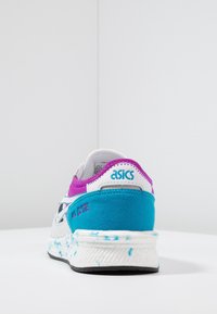 White and teal athletic shoe with purple accents, featuring a suede heel and textured rubber sole. Notable logo and model name visible.