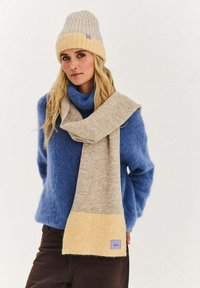 Béžová šála se žlutým spodkem a fialovou nášivkou, nošená s modrým svetrem a ladící béžovou žebrovanou čepicí, vše z měkkých, texturovaných materiálů.
