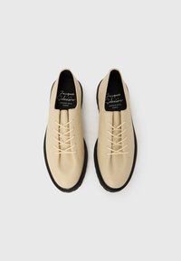 Chaussures en cuir beige à finition texturée, avec un design minimaliste, bout rond, semelles en caoutchouc noir et lacets beiges.