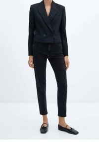 Blazer negro a medida con un diseño de doble botonadura, botones frontales y solapas de pico, combinado con unos pantalones vaqueros negros de corte slim y zapatos planos.