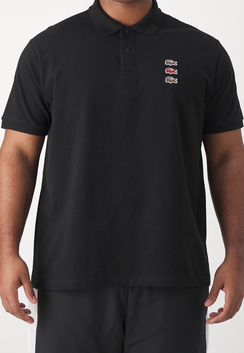 Homme portant un polo noir à manches courtes avec trois petits logos crocodile en brun, rouge et brun sur la poitrine gauche.
