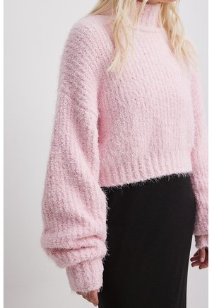 Femme portant un pull en maille rose duveteux avec des manches ballon et une jupe noire sur un fond clair uni.
