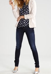 Light gray zip-up jacket over a navy floral blouse, paired with dark blue skinny jeans and shiny silver heels. Simple design and smooth texture.