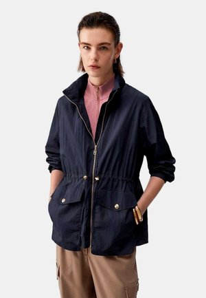 Giovane con capelli corti che indossa una giacca nera con zip sopra una maglietta rosa e pantaloni beige, con le mani nelle tasche, su sfondo neutro.