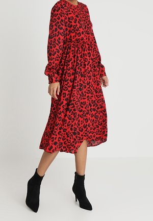 Femme portant une robe midi rouge à imprimé léopard noir, manches longues, et des bottines à talons hauts noires, debout sur un sol gris clair.