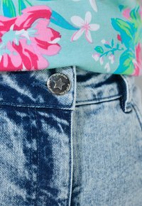 Blå acid-wash denimjeans med en sølvfarvet stjerneformet knap, kombineret med en turkis blomsterprintet skjorte, der er stukket ind i taljen.