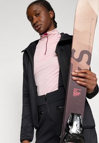 Musta pehmeäkuorinen takki vaaleanpunaista vetoketjupaitaa vasten, mustat housut. Kädessä ruskea suksi mustilla yksityiskohdilla, jossa on graafisia kuvioita.